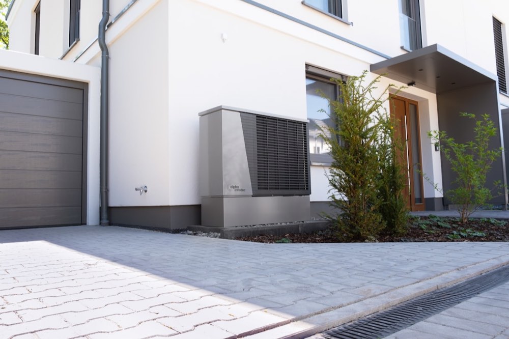 A single grey AC condenser unit installed on the ground beside the exterior wall of a modern home, near the front entrance